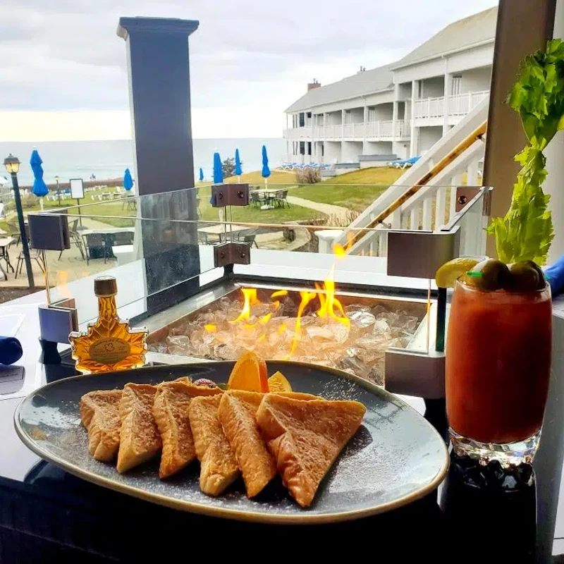 A plate of french toast and a bloody mary sitting on the edge of a fire pit, overlooking the ocean.