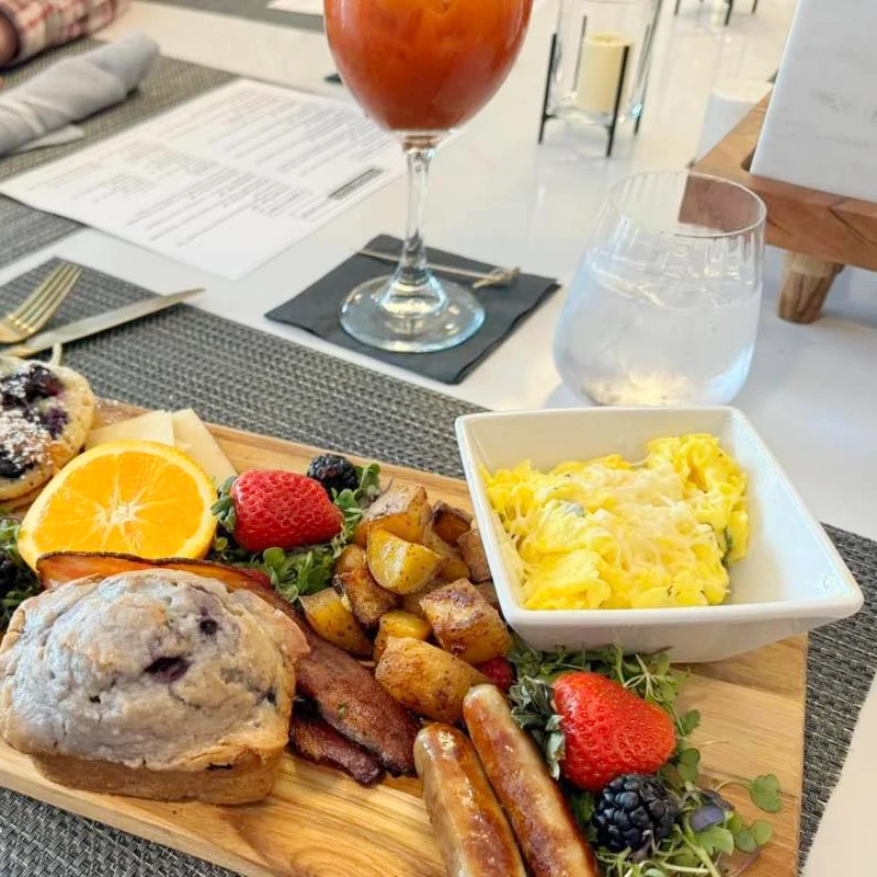 A breakfast spread on top of a wooden plank including eggs, sausage, and a blueberry muffin along with a bloody mary.