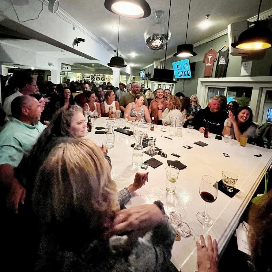 People sitting around a white piano, laughing and talking, with wine glasses and other drinks on top of the piano.